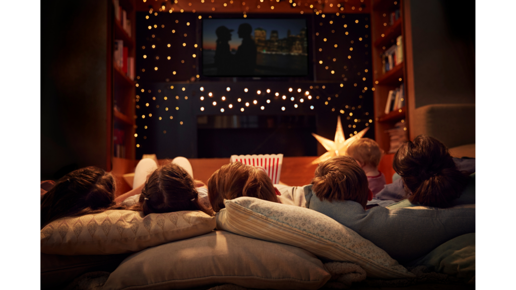 The back of five heads rested on pillows while laying on the ground watching a TV surrounded by fairy lights.